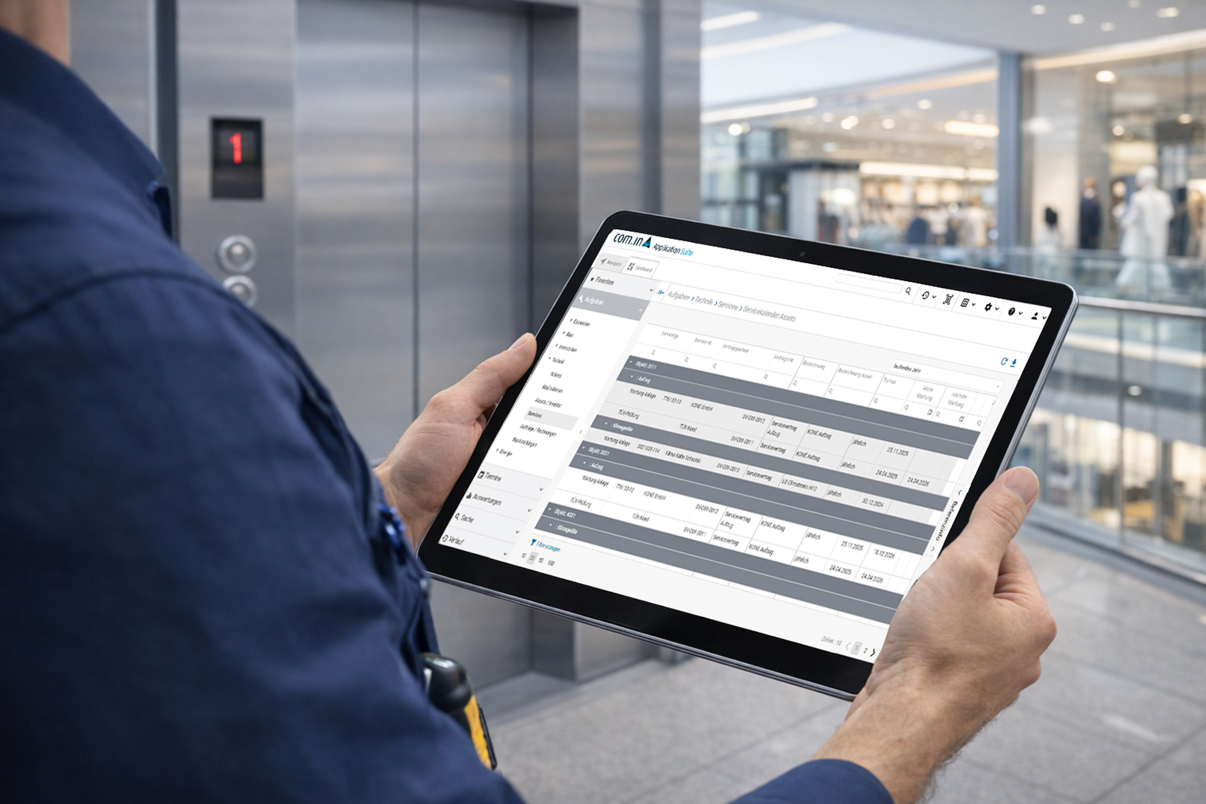 Servicetechniker prüft Wartungsaufgaben auf einem Tablet vor einem Aufzug in einem modernen Einkaufszentrum